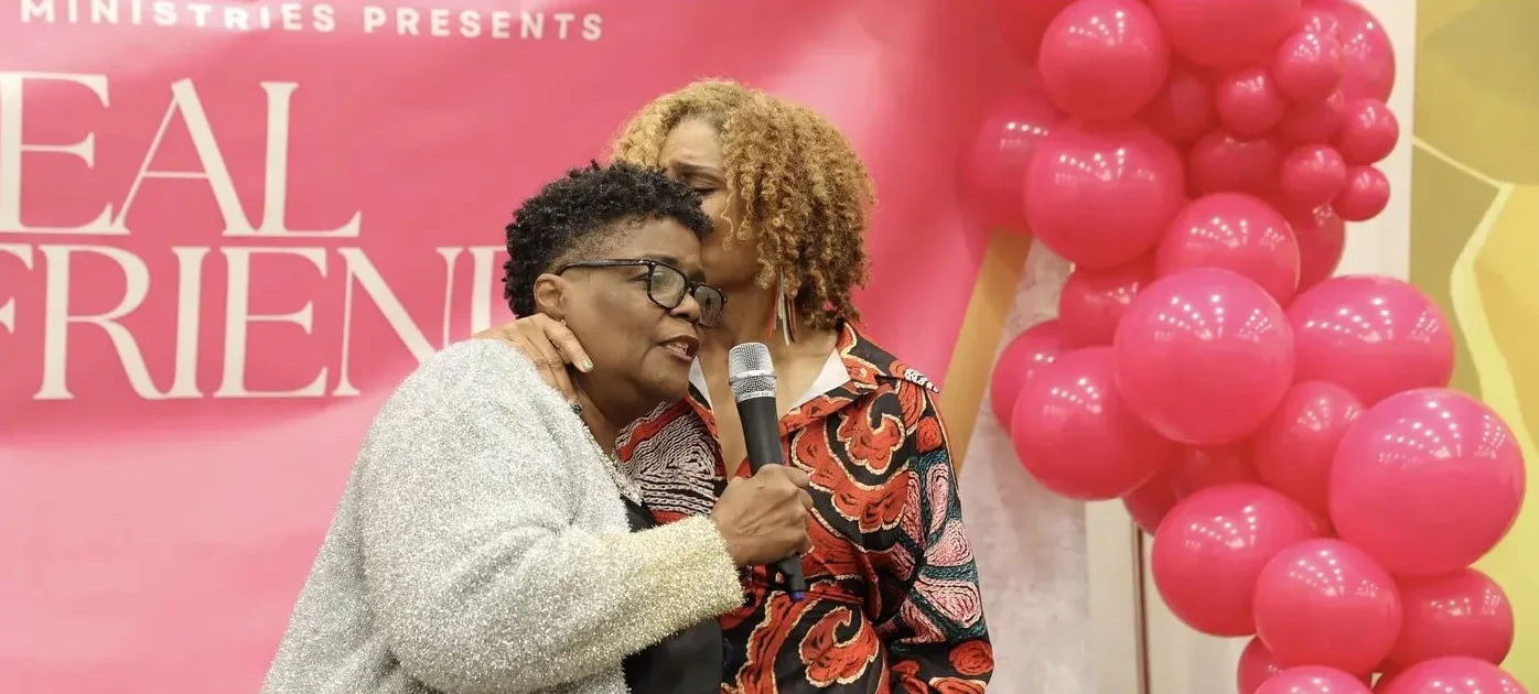 Two women share a warm embrace at the microphone in front of the pink Real Girlfriends banner