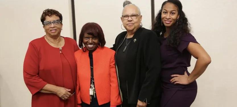 Four women stand together smiling in red, orange, black, and plum