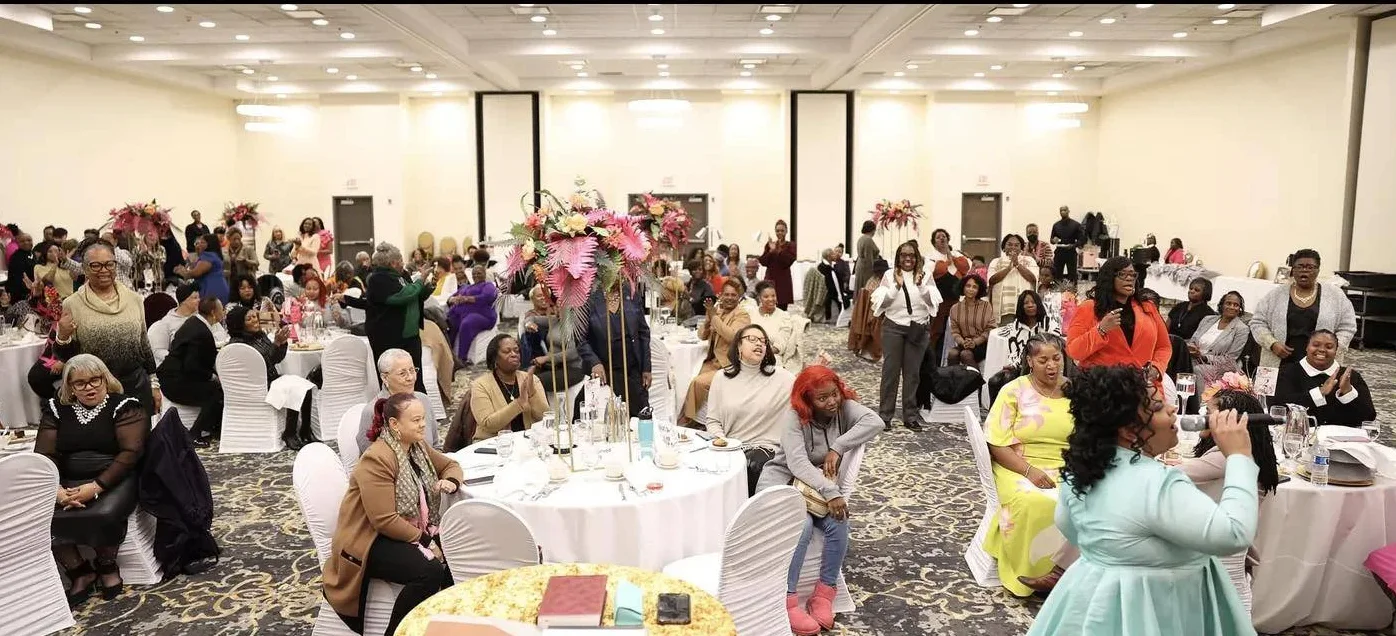A ballroom full of women at round tables turns toward the camera, smiling and waving