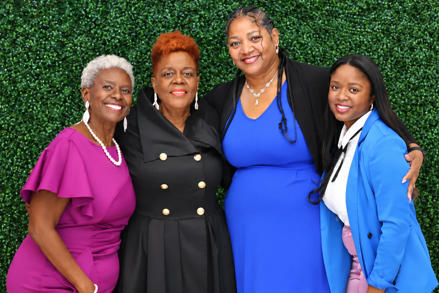 Four women of the Real Girlfriends family stand arm in arm, smiling in front of a greenery wall